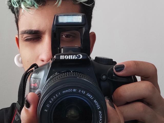 Ezzio, met krullend haar met groene tinten, met één oog gesloten, houdt een zwarte Nikon camera vast, heeft zwarte nagels, witte oordopjes en de lens van de camera is naar voren gericht. Ezzio's gezicht is gedeeltelijk verborgen door de camera..