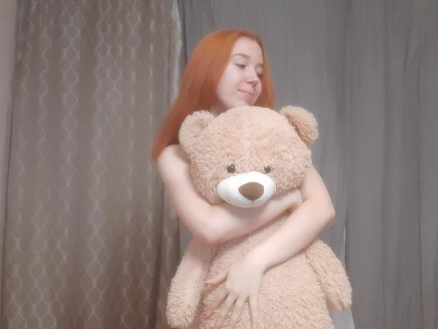 AnikaGlow with red hair hugs a big, fluffy, beige teddy bear against a grey curtain, smiling softly. Her pale skin contrasts with the bear's textured fur.