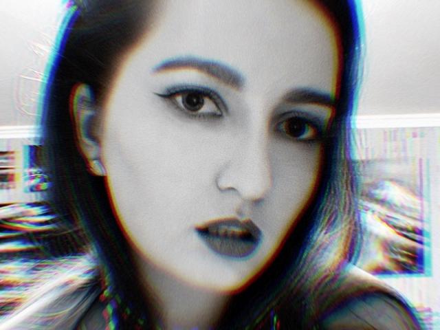 El rostro de EmmyRay, con cabello oscuro y ondulado, sombra de ojos azul y labial azul. Tiene un piercing en la nariz y una ligera sonrisa. La imagen tiene un efecto digital colorido y con fallas..