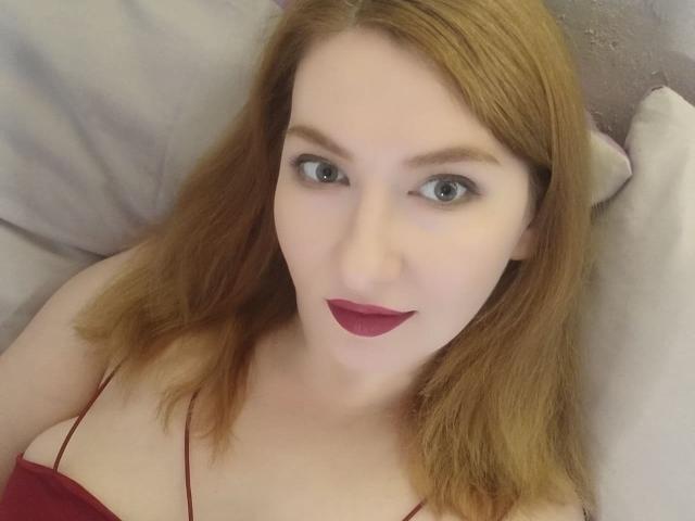 PaulinaSunny with red hair, pale skin, blue eyes, and red lipstick, wearing a red spaghetti strap top, looking up with a slight smile. Boobs visible.