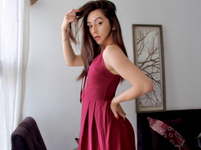LauraMay con un vestito rosa, una mano tra i capelli, che guarda oltre la spalla, con lunghi capelli castani, pelle chiara, trucco leggero, in piedi in una stanza, luce soffusa, divano viola sullo sfondo..