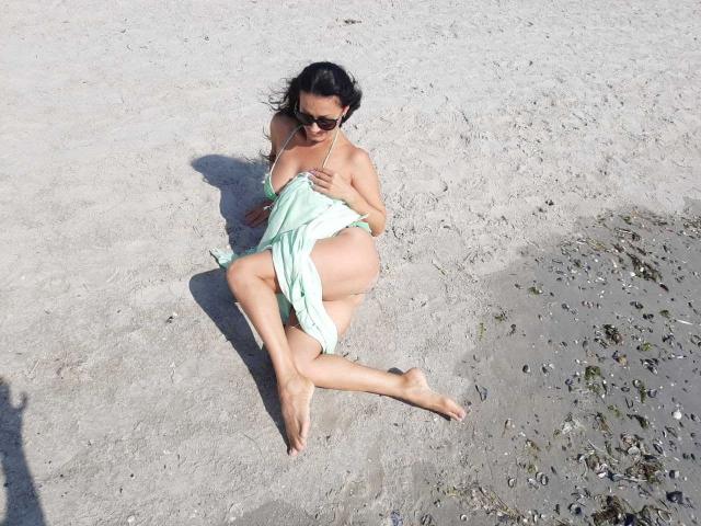 Antralyn, avec ses cheveux foncés et ses lunettes de soleil, assise sur une plage de sable, portant une robe verte, qui expose ses seins et ses fesses, tenant la robe, pieds nus, avec une ombre au sol..