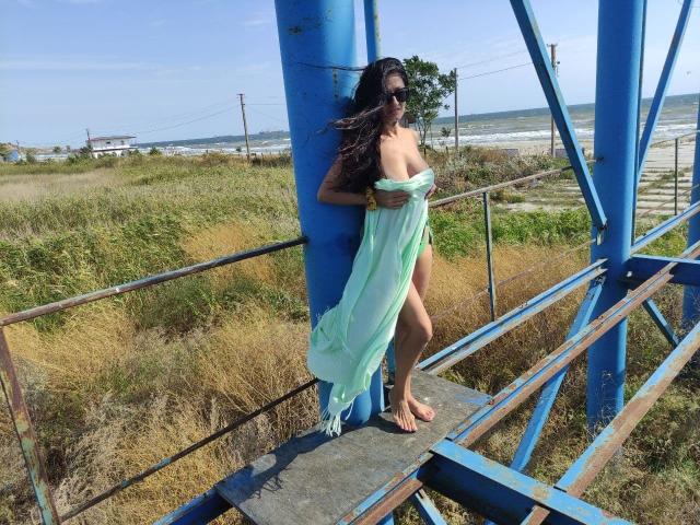 Antralyn with long dark hair, wearing sunglasses, stands on a blue metal structure, covering her tits with a green, sheer, flowing cloth, showing a leg, by the ocean.