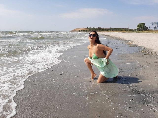Antralyn, agachada em uma praia, vestindo um top de biquíni verde e uma saia transparente, mostrando os seios, com o bumbum levantado, óculos de sol, cabelo escuro e areia molhada..