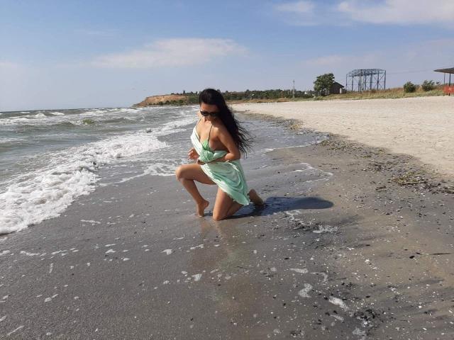 Antralyn knielt op een zandstrand, gekleed in een groene jurk die is opgetrokken om haar blote billen te laten zien, terwijl haar lange zwarte haar in de wind wappert..