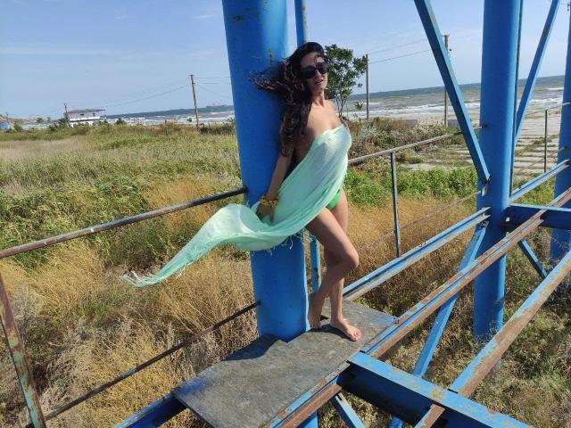 Antralyn with long dark hair, wearing sunglasses, a sheer green scarf, and nothing else, standing barefoot on a blue metal structure, her tits and ass partially visible.