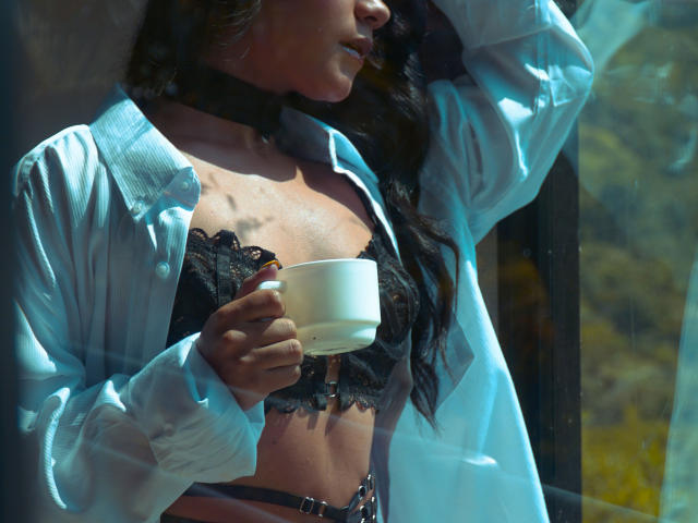AlisonWester met een zwarte kanten bh, wit shirt open, choker, die een witte beker vasthoudt, zonlicht, staand bij het raam, lang donker haar, hand op het hoofd, zwarte riem, zichtbare bruine lijnen, gemiddelde borsten..