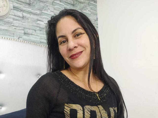 RoraimAnaggie with long black hair, red lipstick, black sheer top, gold necklace, and earrings, smiling at the camera, showing medium tits.
