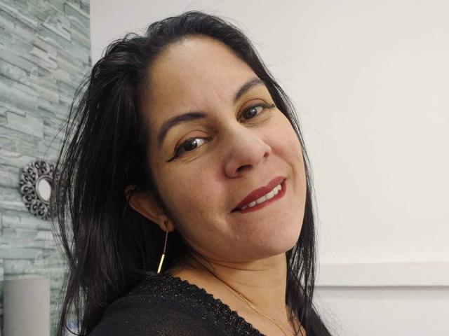 RoraimAnaggie with black hair, brown eyes, red lipstick, smiling, wearing a black top, gold hoop earrings, and a gold necklace. She's got a cute face and a sexy smile.