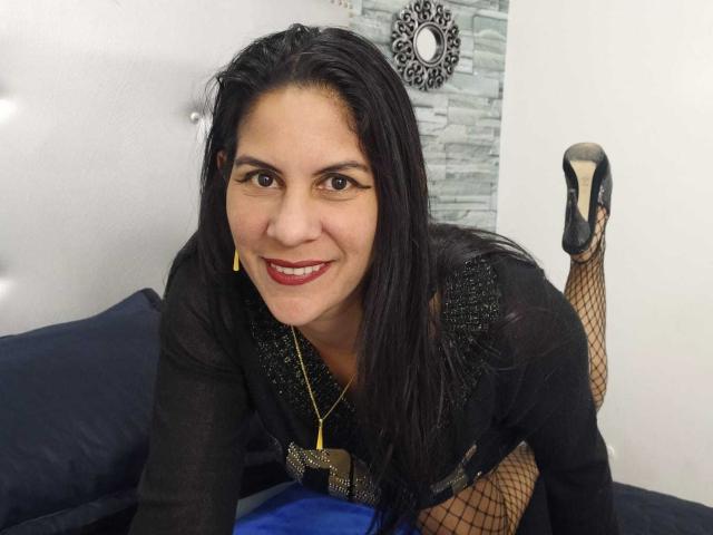 RoraimAnaggie has dark hair, red lipstick, and is smiling while on all fours in a black dress with fishnet stockings and black high heels, showing her ass.