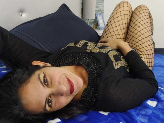 RoraimAnaggie è sdraiata su un letto, indossa un maglione nero, calze a rete e rossetto rosso, con le gambe incrociate, capelli neri e un leggero sorriso, che mostra il sedere..