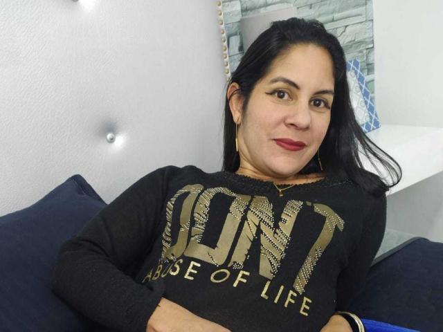 RoraimAnaggie, a Latina woman with black hair, red lipstick, and gold earrings, wearing a black "LUNA" shirt, sitting with arms crossed, smiling subtly.