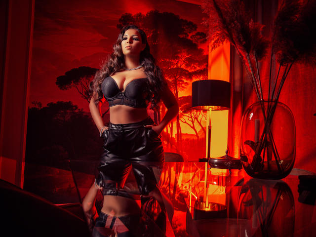 KayaOcean se tient sous un éclairage rouge, vêtue d'un soutien-gorge noir mettant en valeur ses grandes seins et d'une jupe en cuir, les mains dans les poches, avec de longs cheveux ondulés et une expression confiante..