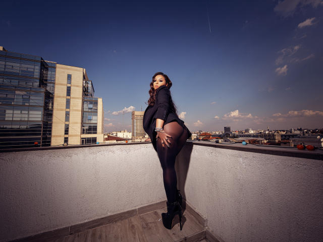 KayaOcean lehnt sich an eine Mauer auf einem Dach, trägt einen schwarzen Blazer, transparente schwarze Strumpfhosen und High Heels, und zeigt ihr Gesäß und ein verführerisches Lächeln, unter einem strahlend blauen Himmel..
