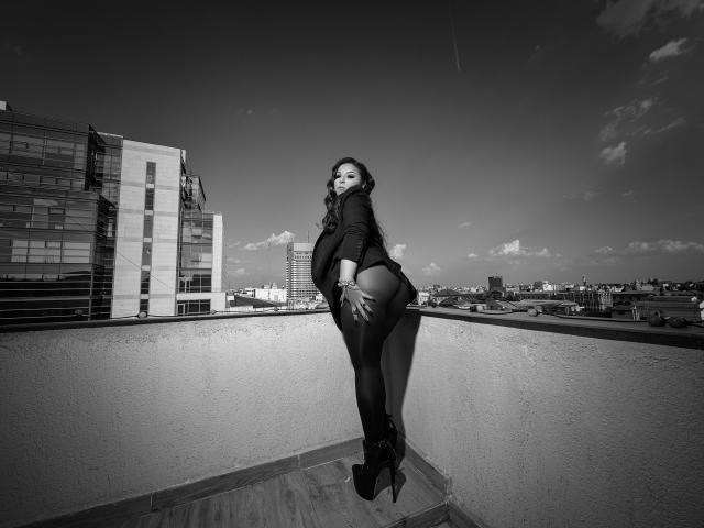KayaOcean, in piedi su un tetto, indossa una maglietta nera, collant neri e tacchi alti, con il sedere in bella mostra, mentre si guarda indietro, in una fotografia in bianco e nero..