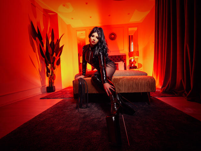 Devasha with long black hair, wearing a shiny black leather outfit and thigh-high boots, kneeling on a bed, holding a black whip, in a red-lit room.