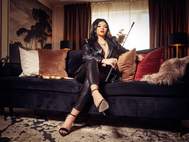 Devasha with black hair, red lipstick, leather jacket, and black pants, sitting on dark blue couch, legs crossed, holding a black strap, wearing high heels, showing her ass.