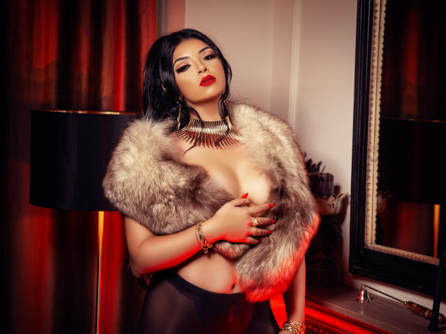 Devasha with black hair, red lipstick, and a fur jacket, covering her tits with one hand, wearing black sheer pants, gold necklace, and bracelet, standing in front of a lamp.