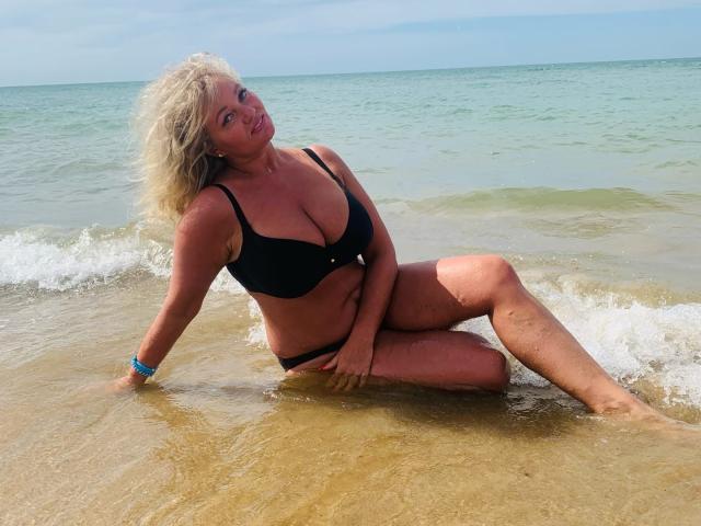 Marmeladka69, bionda, in reggiseno e slip neri, seduta sulla spiaggia, con le gambe immerse nell'acqua, sorridente, con i capelli ricci, pelle abbronzata, braccialetto blu, acqua che tocca le gambe, piccole onde, giornata soleggiata..