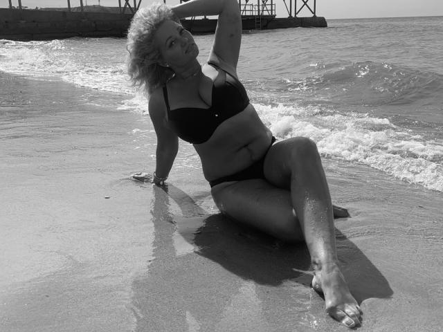 Fotografia in bianco e nero di Marmeladka69 seduta su una spiaggia bagnata, che indossa un bikini scuro, con una mano tra i suoi capelli biondi ricci, mostrando il sedere e i seni..