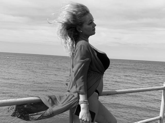 Foto in bianco e nero di Marmeladka69 con lunghi capelli scompigliati dal vento, in piedi su una barca, che indossa un abito largo, mostrando il seno, e braccialetti al polso destro..