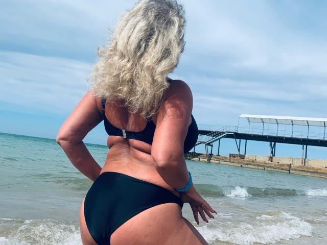Marmeladka69 è in piedi sulla spiaggia, di fronte all'acqua, indossando un bikini nero. Ha i capelli bianchi ricci, la pelle abbronzata e un sedere prosperoso, con la schiena rivolta verso la telecamera..