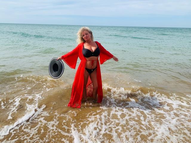 Marmeladka69 è in piedi in acqua bassa, indossa un reggiseno e uno slip neri con una veste rossa, e tiene in mano un cappello. Sono visibili il suo seno grande e i suoi capelli biondi e folti..