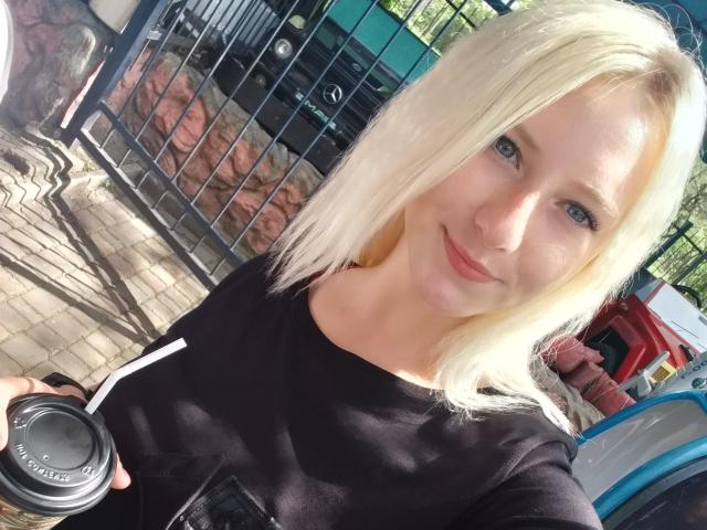 EllenSoNice, avec ses cheveux blonds, ses yeux bleus et son sourire charmant, porte une chemise noire, tient une tasse de café et se tient debout à l'extérieur au soleil, montrant ses fesses sur la photo..