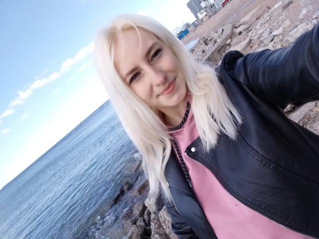 EllenSoNice, met blond haar, lachend, gekleed in een zwarte leren jas over een roze shirt, staand bij de rotsachtige kust, met een blauwe oceaan op de achtergrond..