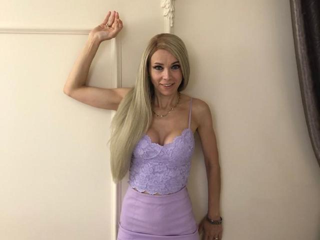 Kassandra Gray, con lunghi capelli biondi, indossa una blusa e una gonna di pizzo color lavanda, è in piedi contro un muro, sorride, con un braccio alzato, seno piccolo, figura esile, pelle chiara, collana dorata..