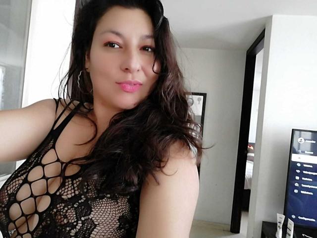 SakotoDika with wavy dark hair, pink lipstick, wearing a black netted top showing her boobs, hoop earrings, in a white room, looking at the camera with a slight smile.