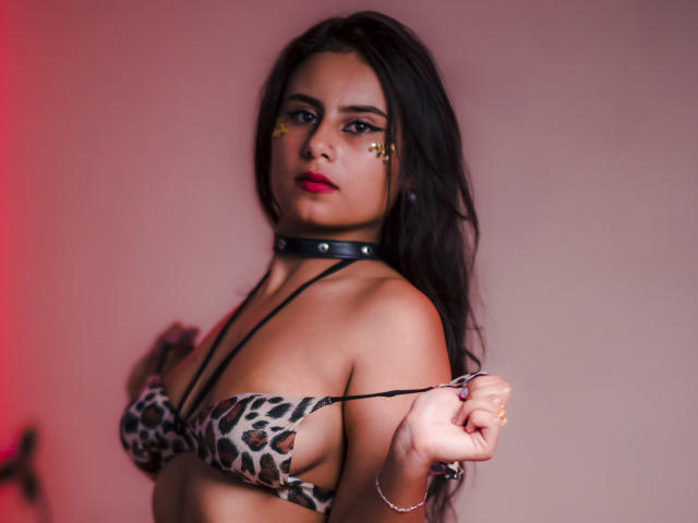 LeilaEvanns mit langen, schwarzen Haaren, die einen BH mit Leopardenmuster, eine schwarze Halskette und goldene Sternaufkleber auf ihren Wangen trägt, zieht an einem BH-Bügel und zeigt ihre Brüste. Rotes Licht..