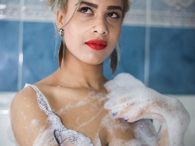 Kendall Quinn avec des bulles de savon sur ses seins, du rouge à lèvres, et des boucles d'oreilles bleues, regardant vers le haut avec un léger sourire, les cheveux blonds en un chignon désordonné, dans une salle de bain..