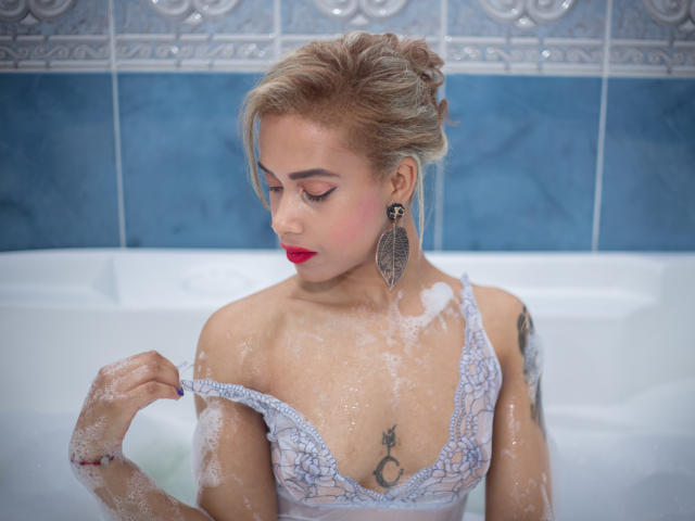 KendallQuinn with wet, soapy skin, lifting a lace-covered tit, red lipstick, blonde updo, tattoos, large earrings, in a blue-tiled bathroom, looking down, showing a sexy, confident vibe.