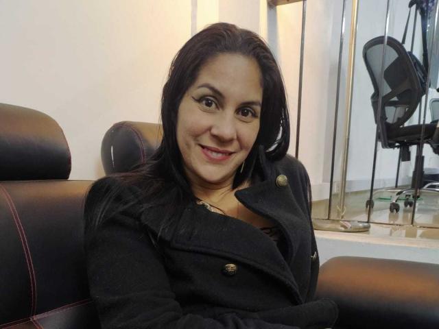 RoraimAnaggie has dark hair, brown eyes, and a cute smile, wearing a black coat with buttons, sitting in a black leather chair, looking happy and relaxed.