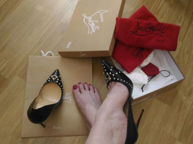 AlfaFemale's feet with red-painted toenails wearing black studded heels, next to brown boxes and a red La Perla bag on a wooden floor.