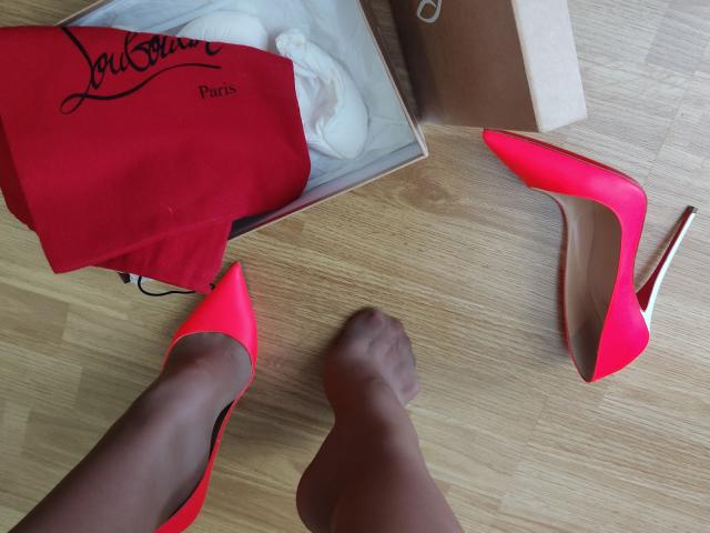AlfaFemale's legs in sheer black pantyhose with bright red pointed heels, one shoe on and one off, near a red bag and white box on a wooden floor.