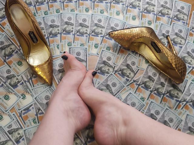 AlfaFemale's feet with black-painted toenails, crossed at the ankles, above two golden high heels and a pile of money on the floor.