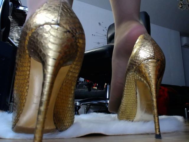 Gold snake-skin high heels with tall stiletto heels, worn by AlfaFemale, standing on a white furry rug, showing her bare feet and legs.