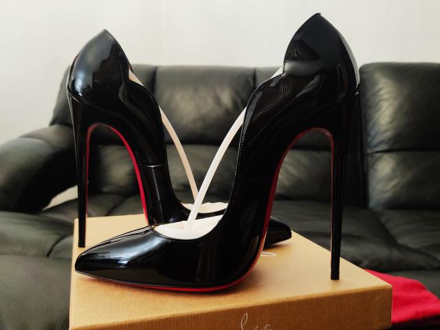Glossy black stiletto heels with red soles, standing on a cardboard box, straps crossed in the middle, on a black leather couch. The heels are a bitch.