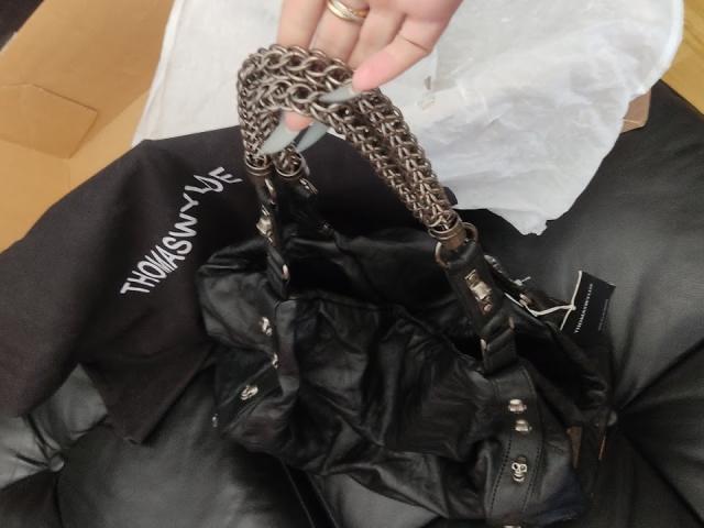 AlfaFemale's hand holding a black leather bag with silver chain handles, sitting on her black jeans-covered ass. Bag's straps and zippers are visible.