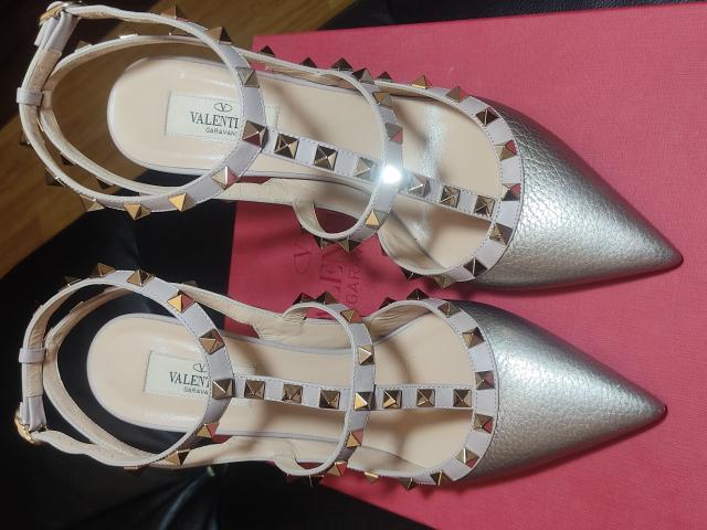 A pair of shiny, silver, pointed-toe Valenti heels with white straps and gold spikes, placed on a pink surface. The soles are beige. AlfaFemale's shoes.
