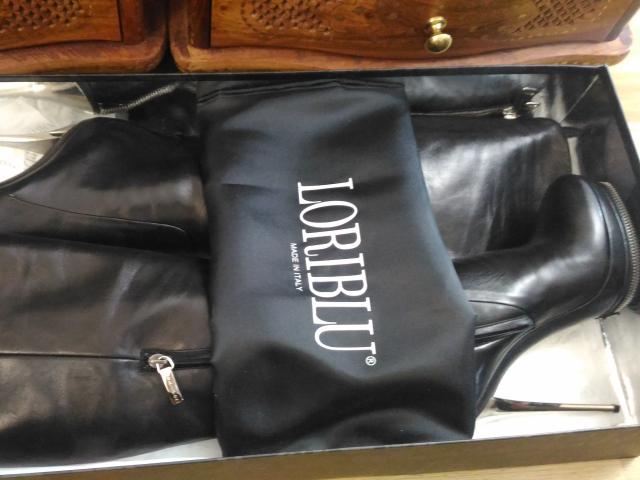 Black boots with shiny leather, zipped sides, and a black bag with "Dior" written on it, placed inside a box. Boot bag covers the boots' ass.