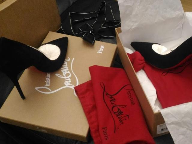 Black stiletto heels with white soles, one shoe on a brown box, the other on a red cloth, and a black dress with white trim in the background.