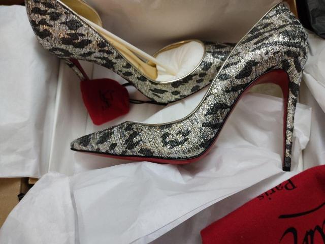 Une paire d'escarpins hauts brillants, argentés et noirs avec un motif serpent, et des semelles rouges, reposant sur un tissu blanc, à côté d'un sac en velours rouge. Les escarpins sont posés sur une feuille de papier blanche..