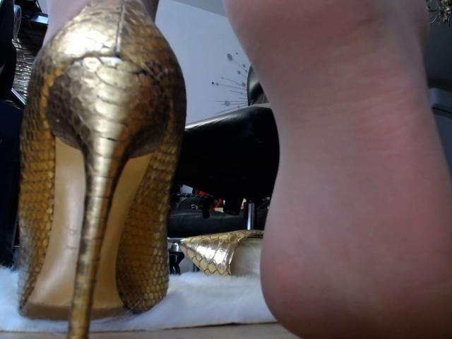 Gold stiletto heels with snake skin pattern, foot with pinkish sole, AlfaFemale's heel up, white furry surface, high heel on left, visible sole on right.