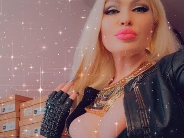 AlfaFemale with blonde hair, pink lipstick, black leather jacket, gold necklace, and black fingerless gloves, showing her big tits, with sparkles in the photo.