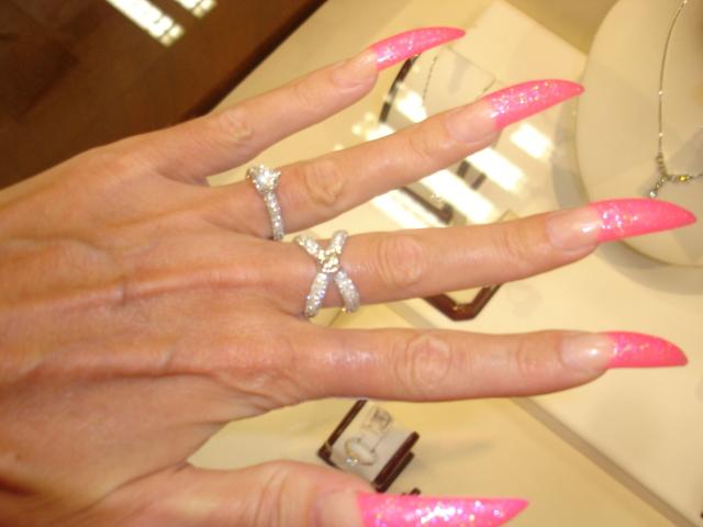 AlfaFemale's hand with pink glitter nails, wearing three diamond rings on her fingers, in a bright store with jewelry displays. Her fingers are slightly spread, showcasing her nails.
