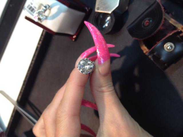A unha rosa com glitter da AlfaFemale, com um grande diamante brilhante no dedo indicador, ao lado de suas unhas longas e pontudas cor de rosa, com uma bolsa de câmera preta ao fundo..
