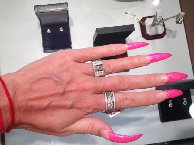 La main d'AlfaFemale, avec des ongles roses pailletés, portant deux bagues en argent avec des diamants, un bracelet rouge, une boîte de présentation noire, et un porte-clés avec des lunettes en arrière-plan..